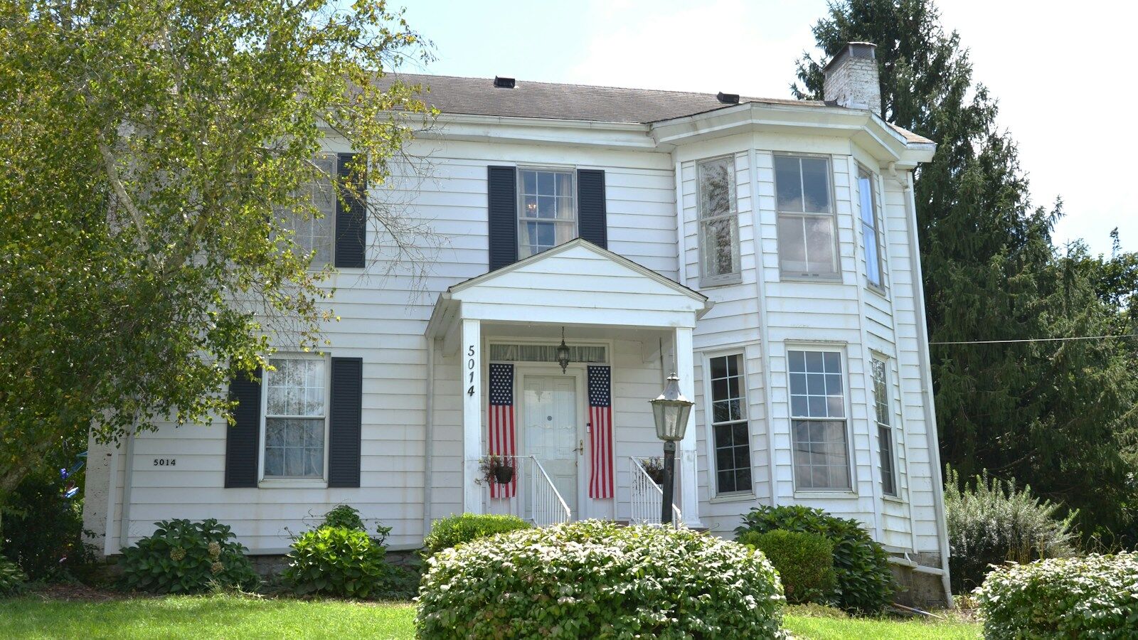 A white two-storey house with black shutters and mature trees on a residential street.